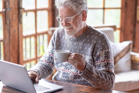 Ein älterer Mann mit einer Kaffeetasse in der Hand und einem Laptop, auf der Suche nach einer Immobilienbewertung